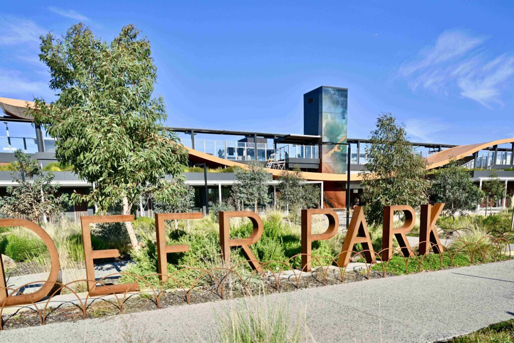 The distinctive rusted metal "DEER PARK" sign set within a native garden, with the modern station architecture and stone paving from Eigen Stones in the background.