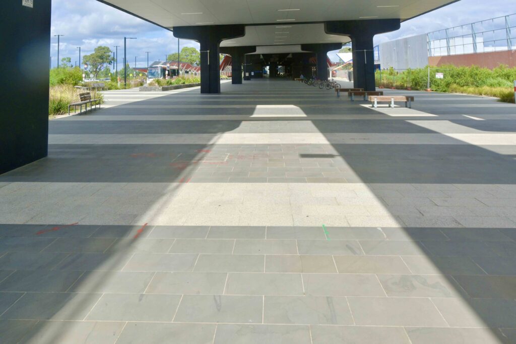 The vast, sheltered space beneath the Mernda Station rail bridge, paved with a striped pattern of light and dark stone from Eigen Stones.